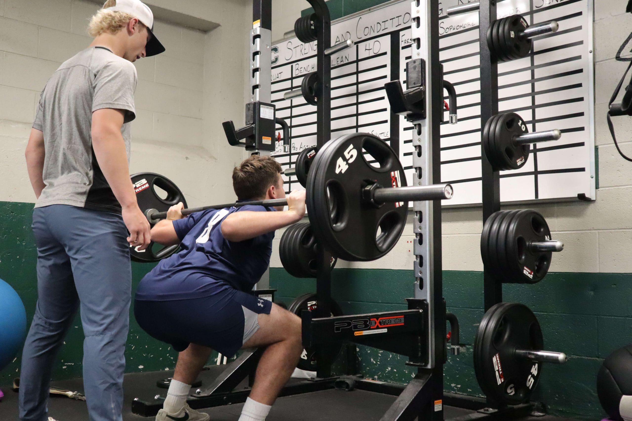 A student performs the barbell back squat in a high school weight room.
