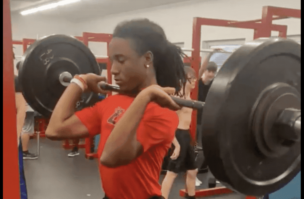 A student holds a barbell in the front rack during a high school strength and conditioning physical education class.