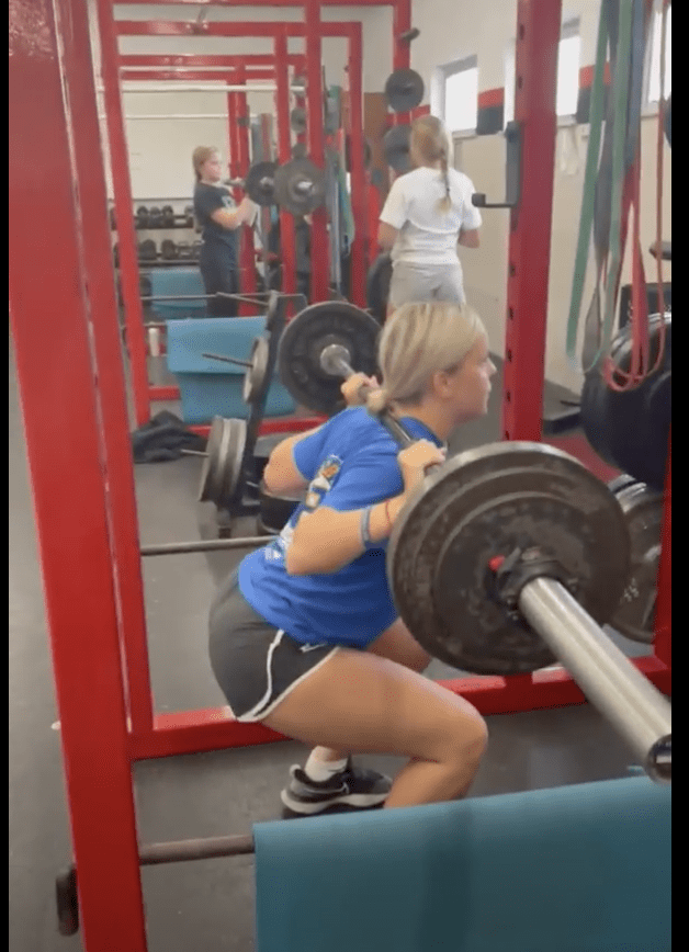 A student performs a barbell back squat in a strength and conditioning physical education class.